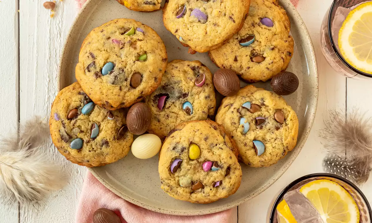 Cookies dorés parsemés de pépites colorées et morceaux de chocolat, posés sur une assiette beige entourée de décorations de Pâques