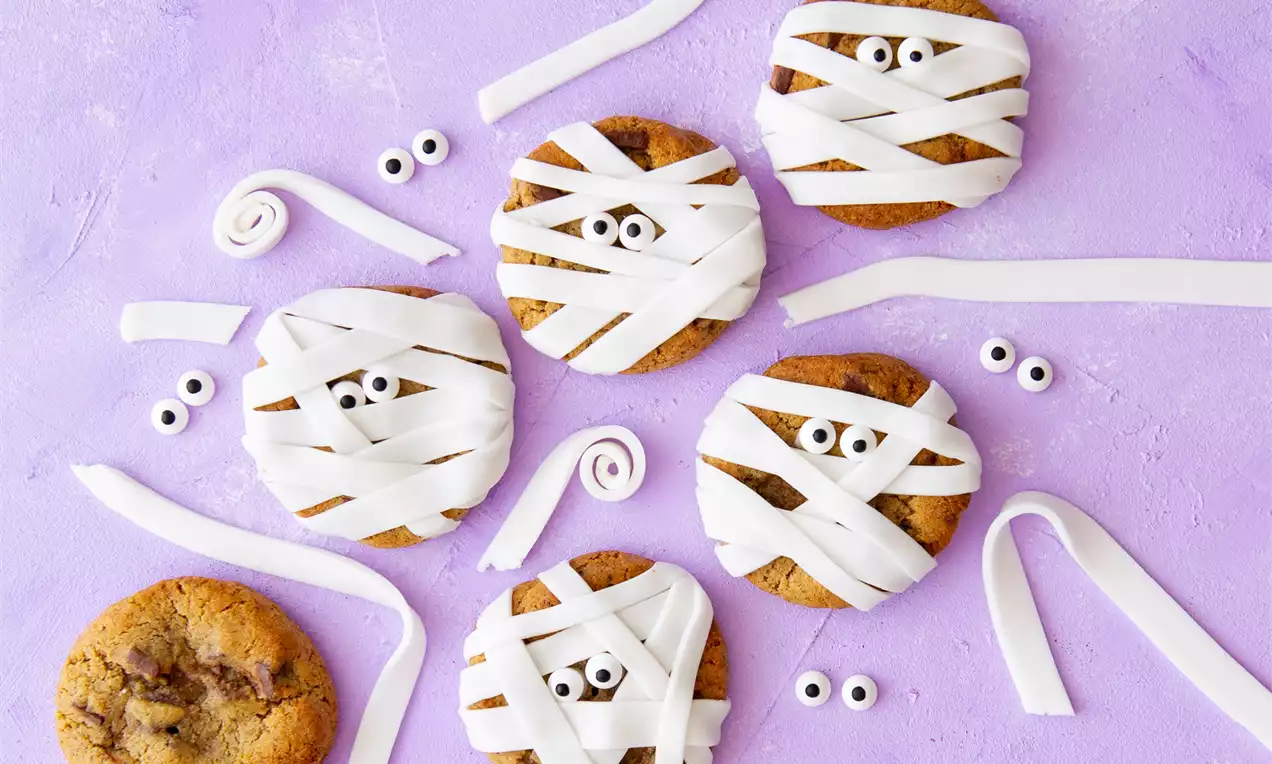 Bruingebakken ronde cookies met witte fondantbanen als wikkels, versierd met zwarte oogjes op een lila achtergrond met losse fondantstroken
