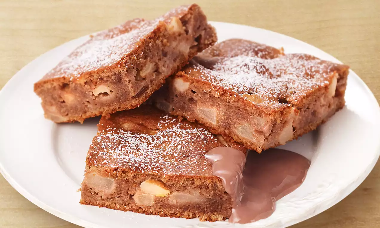 Carrés moelleux de gâteau aux pommes et aux noisettes dorés, saupoudrés de sucre glace, disposés sur une assiette blanche avec une touche de sauce chocolatée
