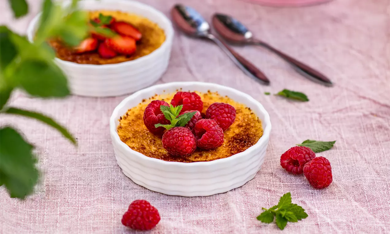 Крем Брюле с карамелизирана златиста коричка, декориран с пресни малинови плодове и ментови листа в бяла керамична купичка