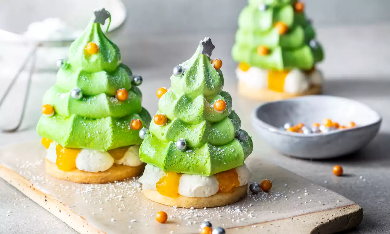 Petits sapins vert vif en meringue ornés de billes colorées et étoiles, posés sur une planche beige avec des éclats de sucre