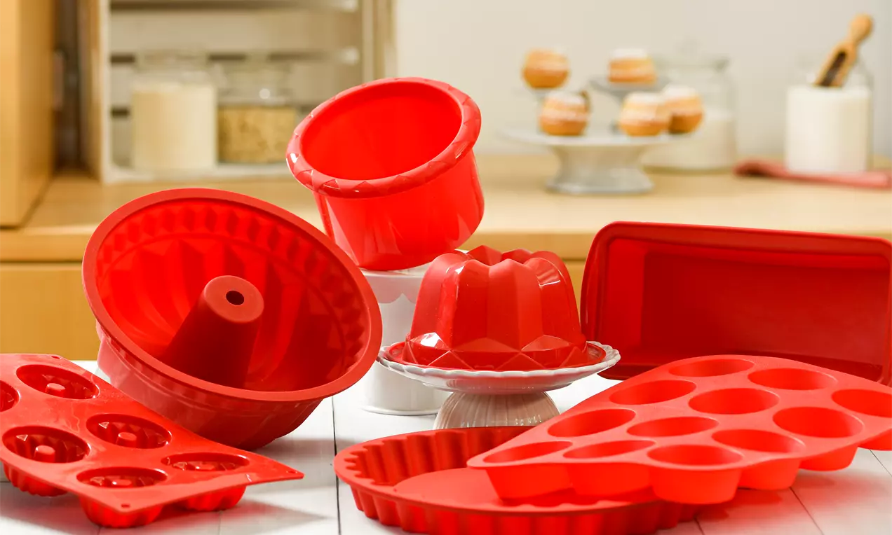 Червени силиконови форми с различни очертания и детайли, разположени върху бял дървен плот в кухненска обстановка с топли акценти