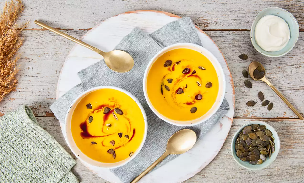 Hokkaido-Kürbissuppe leuchtend orange mit cremiger Textur, garniert mit Kürbiskernen und dunklen Tropfen in weißen Schalen auf hellem Holztablett