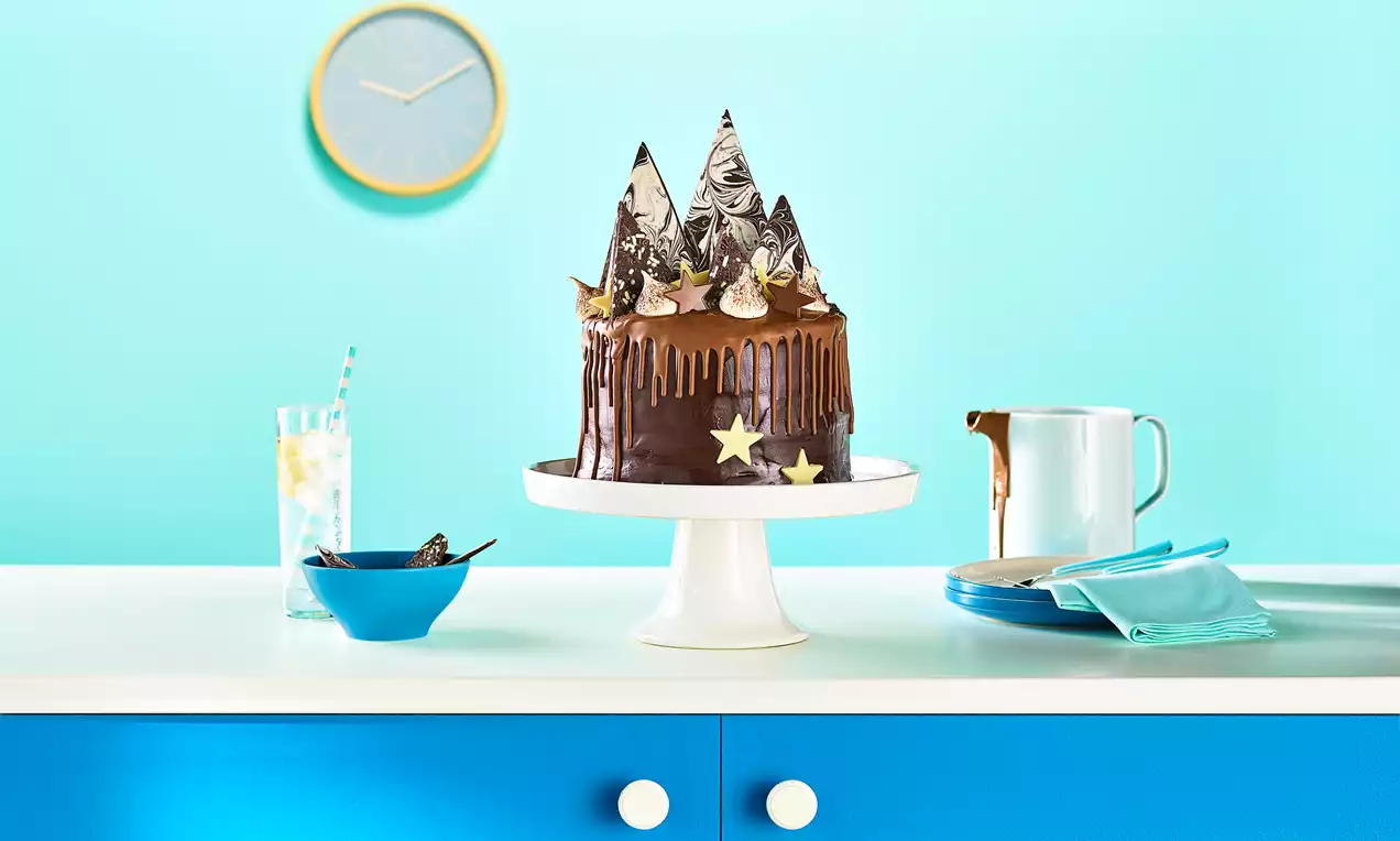 Chocolate cake with glossy ganache drips, pointed chocolate shards, cream swirls, and star decorations on a white cake stand