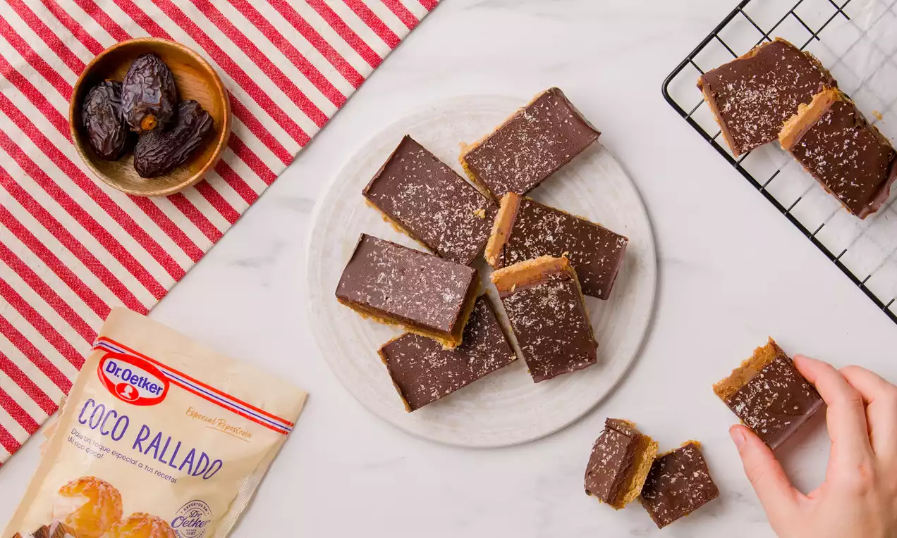 Barritas rectangulares con cubierta de chocolate oscuro espolvoreado con coco rallado sobre base dorada de caramelo dispuestas en plato blanco