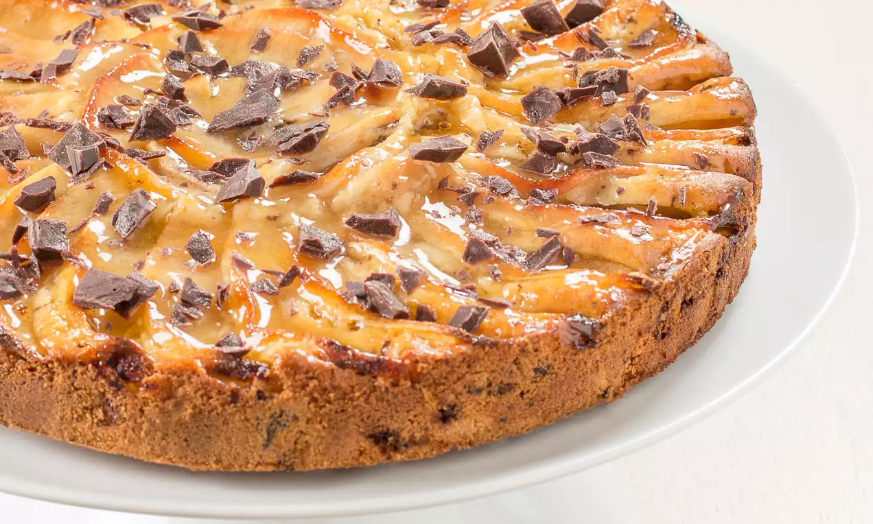 Torta dorata con fettine di mela caramellate disposte a raggiera, punteggiata da scaglie di cioccolato su un piatto bianco