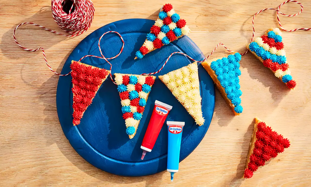 Colourful triangular blondie pieces arranged like bunting on a blue plate, decorated with piped red, blue and yellow icing rosettes