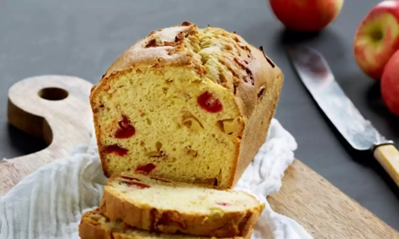 Avlang julekake med gyllenbrun skorpe, røde fruktbiter og eplebiter, delvis skåret på trefjøl med hvitt klede og røde epler ved siden av