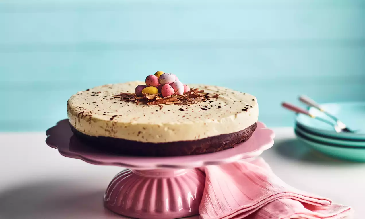 Rund ostekake med lys kremfarget fyll på mørk bunn, pyntet med sjokoladeskav og små fargerike påskeegg på rosa kakefat