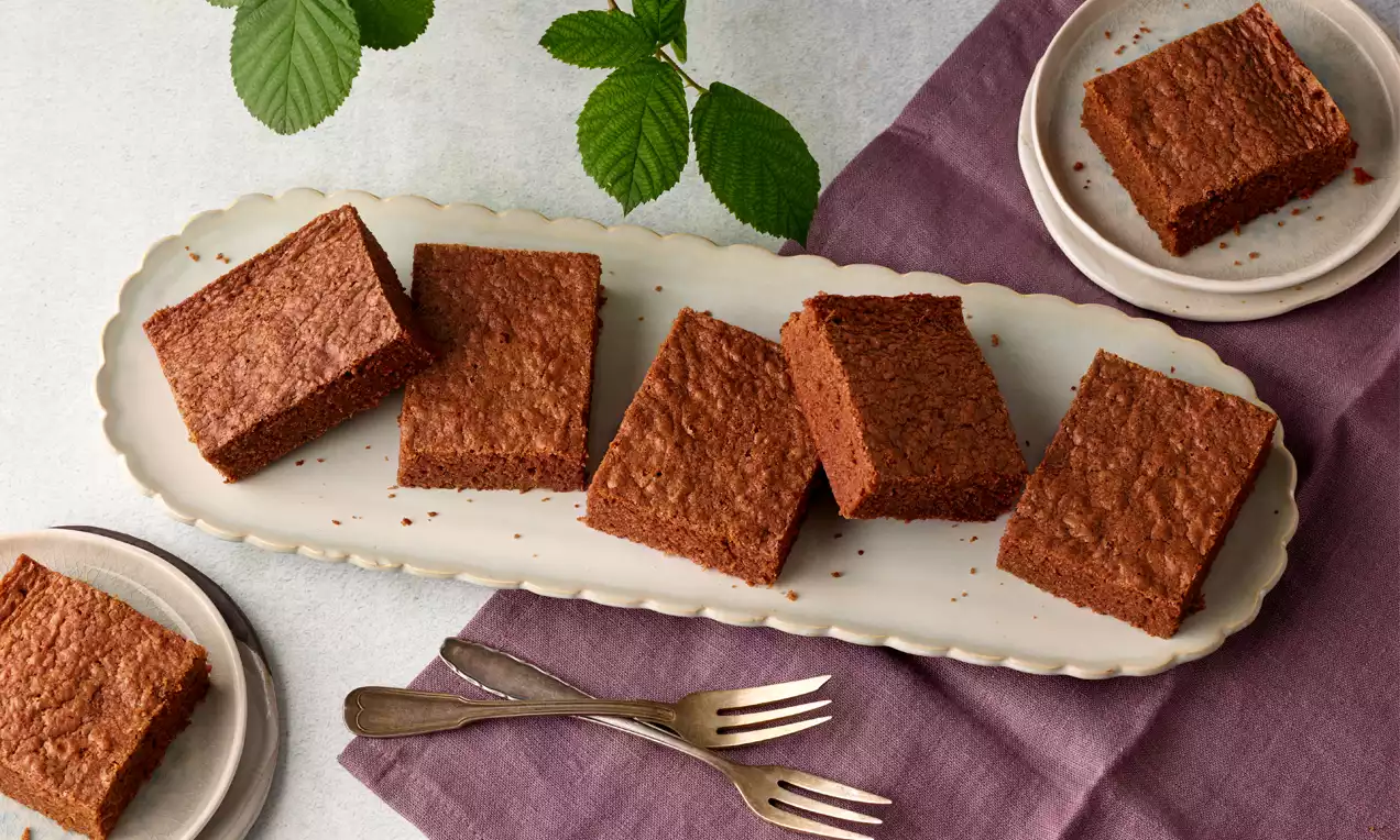Rechteckige, schokoladenbraune Brownies mit saftiger Textur auf heller Keramikplatte, dekoriert mit lilafarbener Stoffserviette und Kräuterzweigen