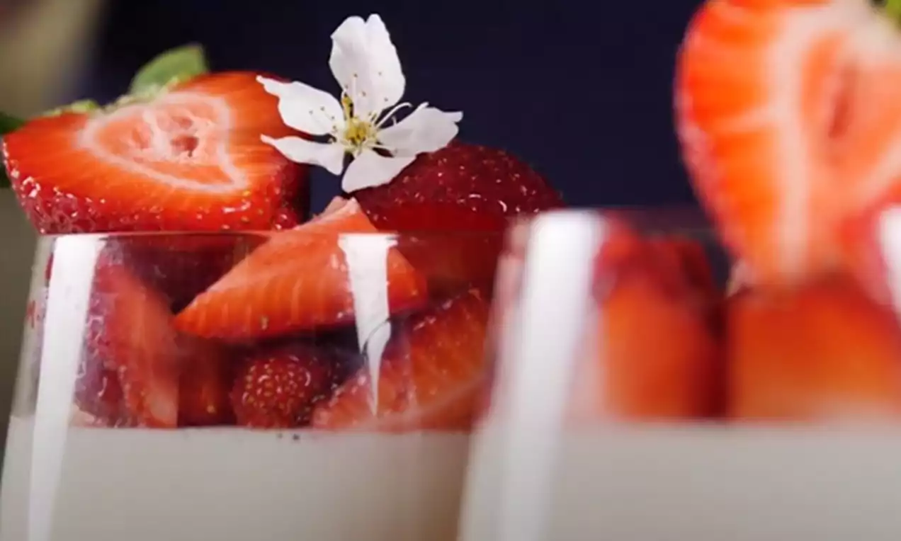 Kristāldzidra zemeņu želeja, maigi baltā Panna Cotta, glāzē ar sulīgu zemeņu gabaliņiem un baltu ziedu akcentu uz virsmas