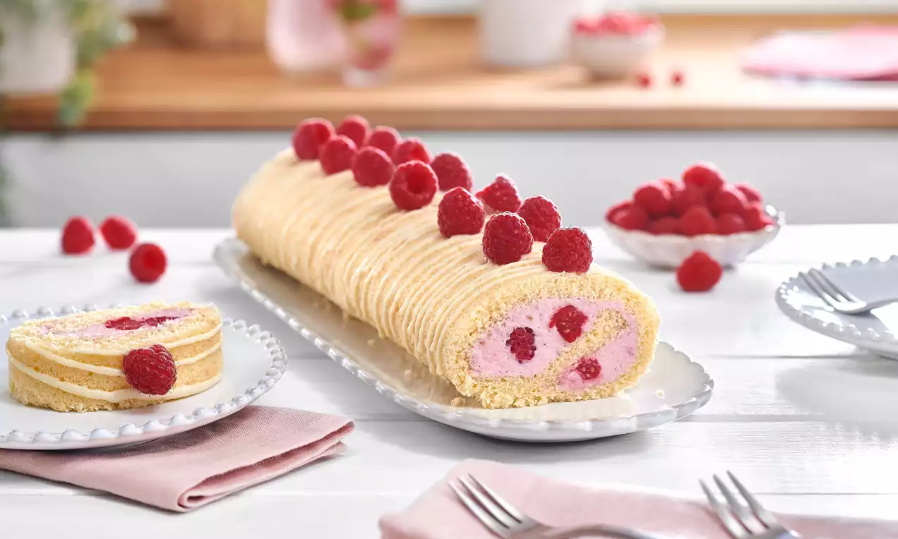 Helle, goldgelbe Biskuitrolle mit rosa Himbeer-Joghurt-Füllung, frischen Himbeeren und gewelltem Muster auf länglicher Silberplatte