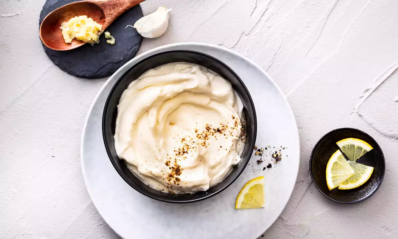 Salsa alioli cremosa de color marfil en cuenco negro sobre plato gris claro, decorada con pimienta y rodajas de limón