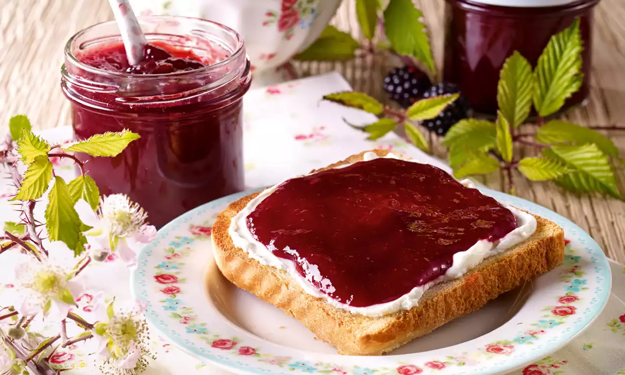 Fetta di pane dorata spalmata di confettura rosso scuro lucida su crema bianca, accanto a un barattolo colmo su tovaglietta floreale