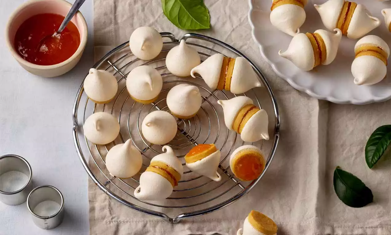 Kleine Zwickerbusserln aus weißem Baiser mit goldgelber Füllung aus Aprikosenmarmelade auf einem silberfarbenen Kuchengitter drapiert