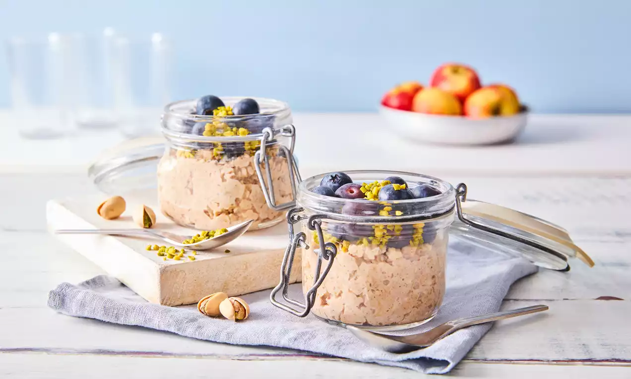 Zwei Gläser mit cremigen Overnight-Oats, getoppt mit blauen Beeren und gelben Pistaziensplittern, auf einem hellen Holzbrett mit Deko