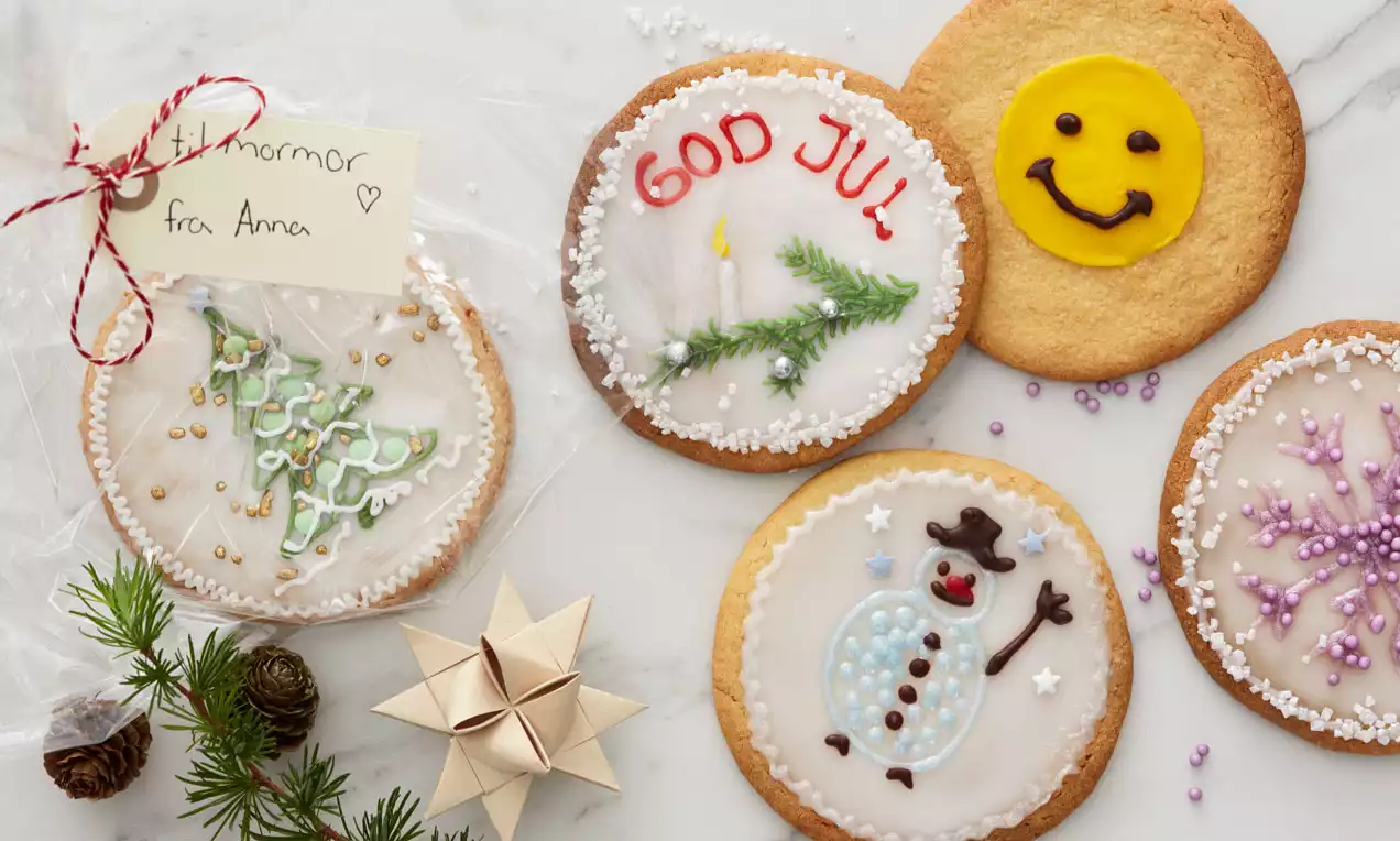Store, runde småkaker med hvit glasur dekorert med julemotiv som tre, snømann og tekst, plassert på hvit marmor med granbar og stjernefletting