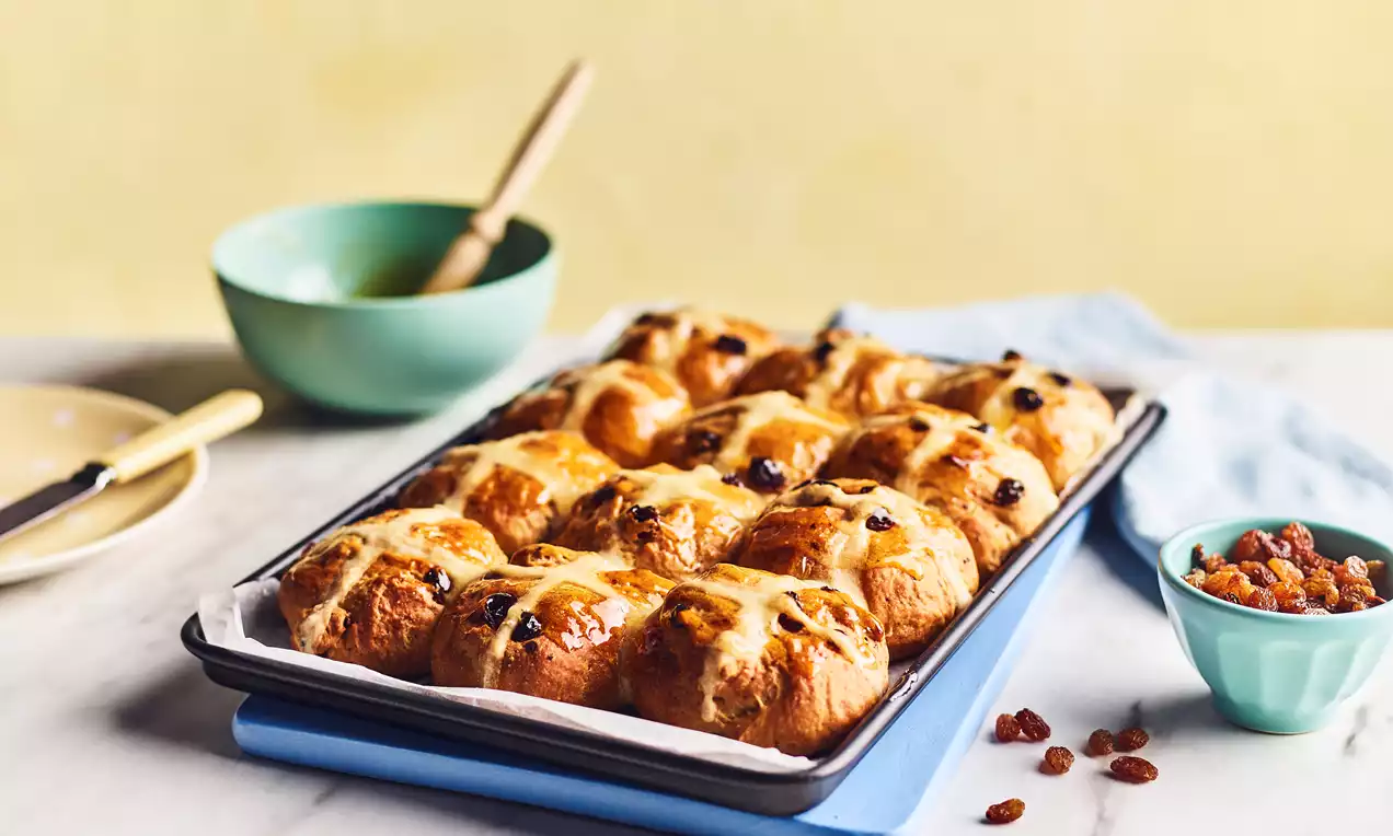 Golden-brown vegan hot cross buns with glossy glaze and white flour crosses arranged in rows on a baking tray with scattered raisins nearby