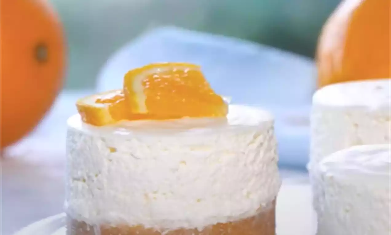 Majhen okrogel cheesecake z zlato rjavo podlago, puhasto belo kremo in krhljem živo oranžne pomaranče na vrhu