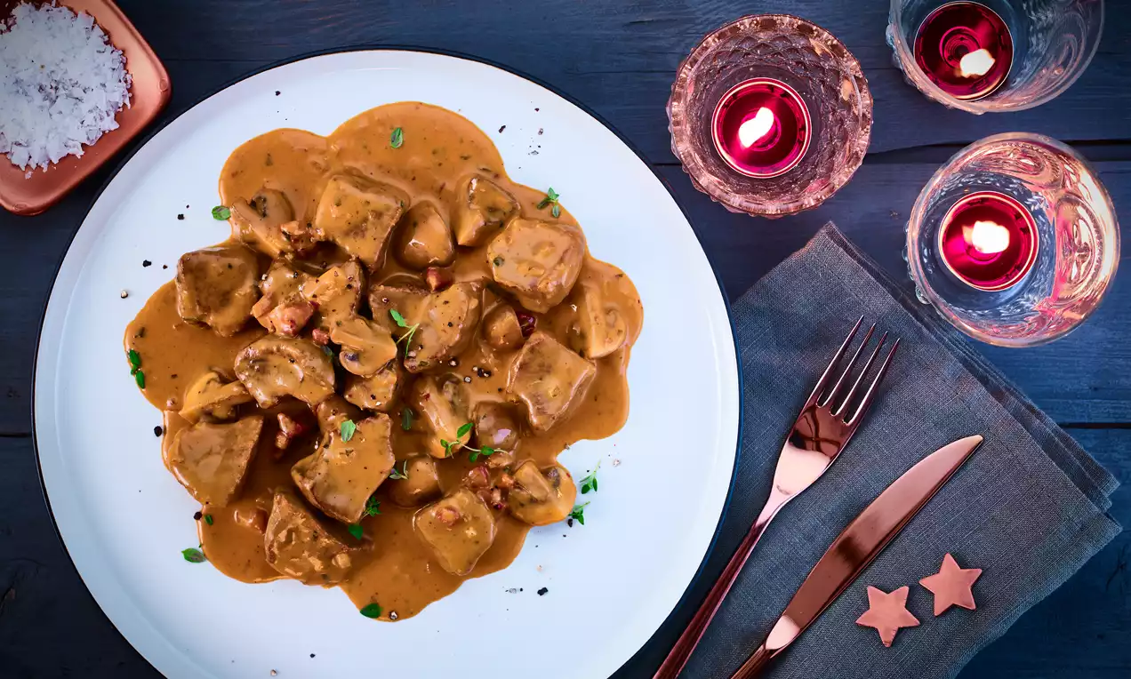 Hirschgulasch in zartbrauner, cremiger Sauce mit Kräutern garniert auf weißem Teller, arrangiert auf dunkler Holzoberfläche mit Kerzendekoration