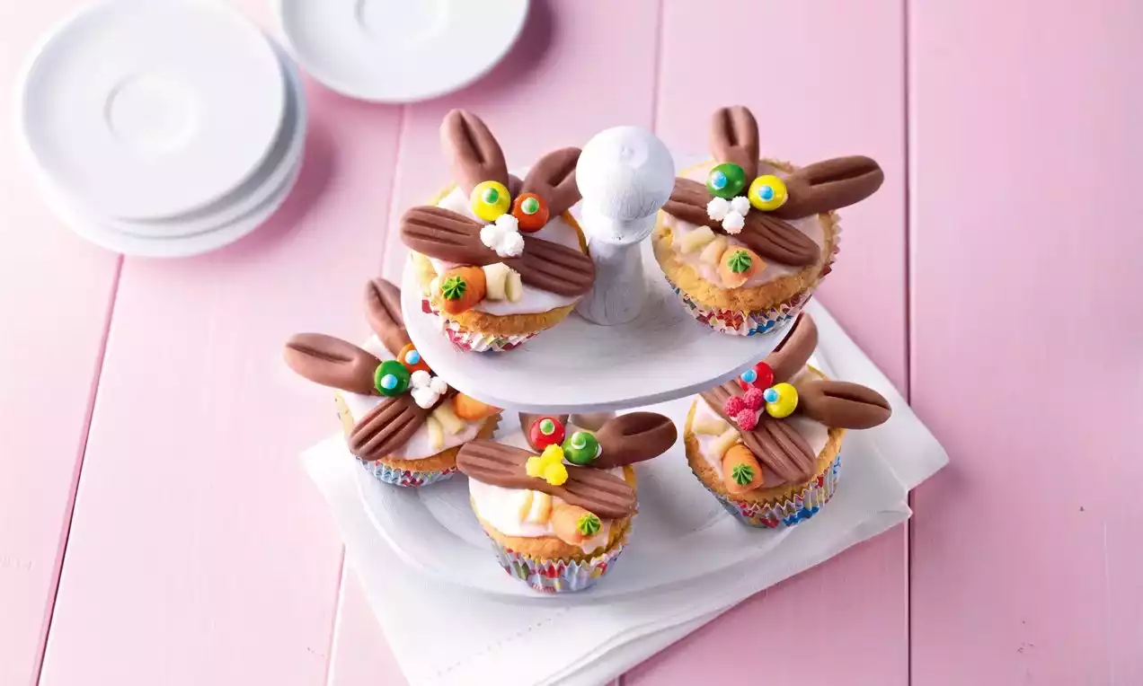 Easter Bunny cupcakes with colorful icing, chocolate ears, candy decorations, on a white tiered stand against a pink wooden surface