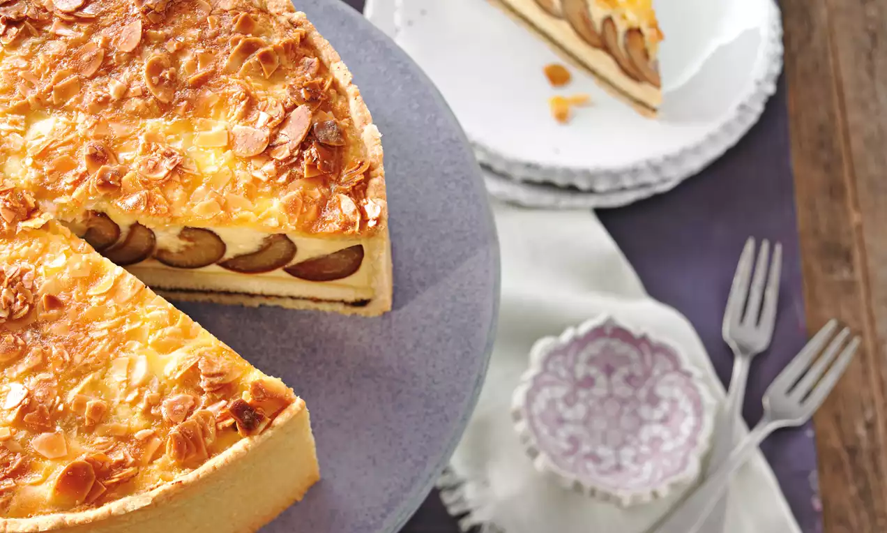 Okrugli kolač s prhkom svijetlom podlogom, zlatnom kremom prekrivenom listićima badema i slojem šljiva, poslužen na platou