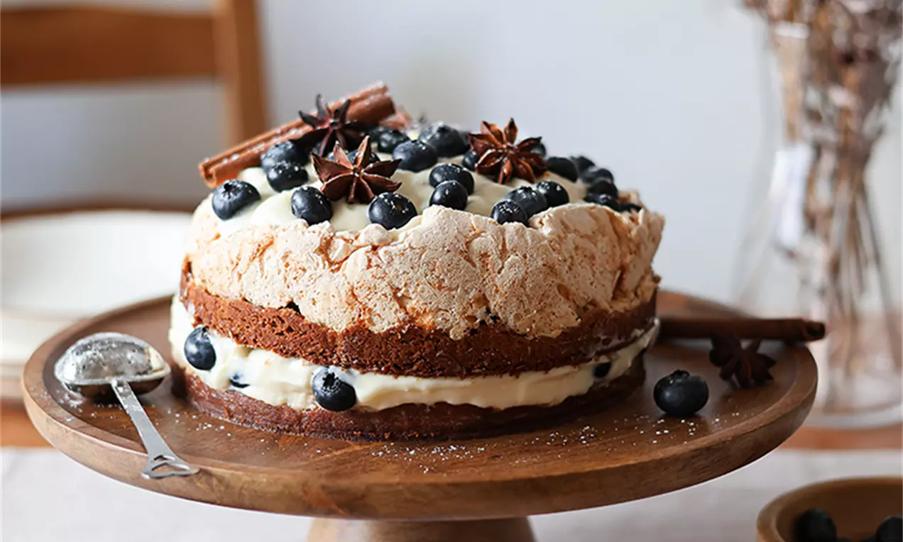 Слоеста торта с кафяв блат, пухкав крем и хрупкава меренга, покрита с пресни боровинки, звездовиден анасон и канелени пръчици върху дървена стойка