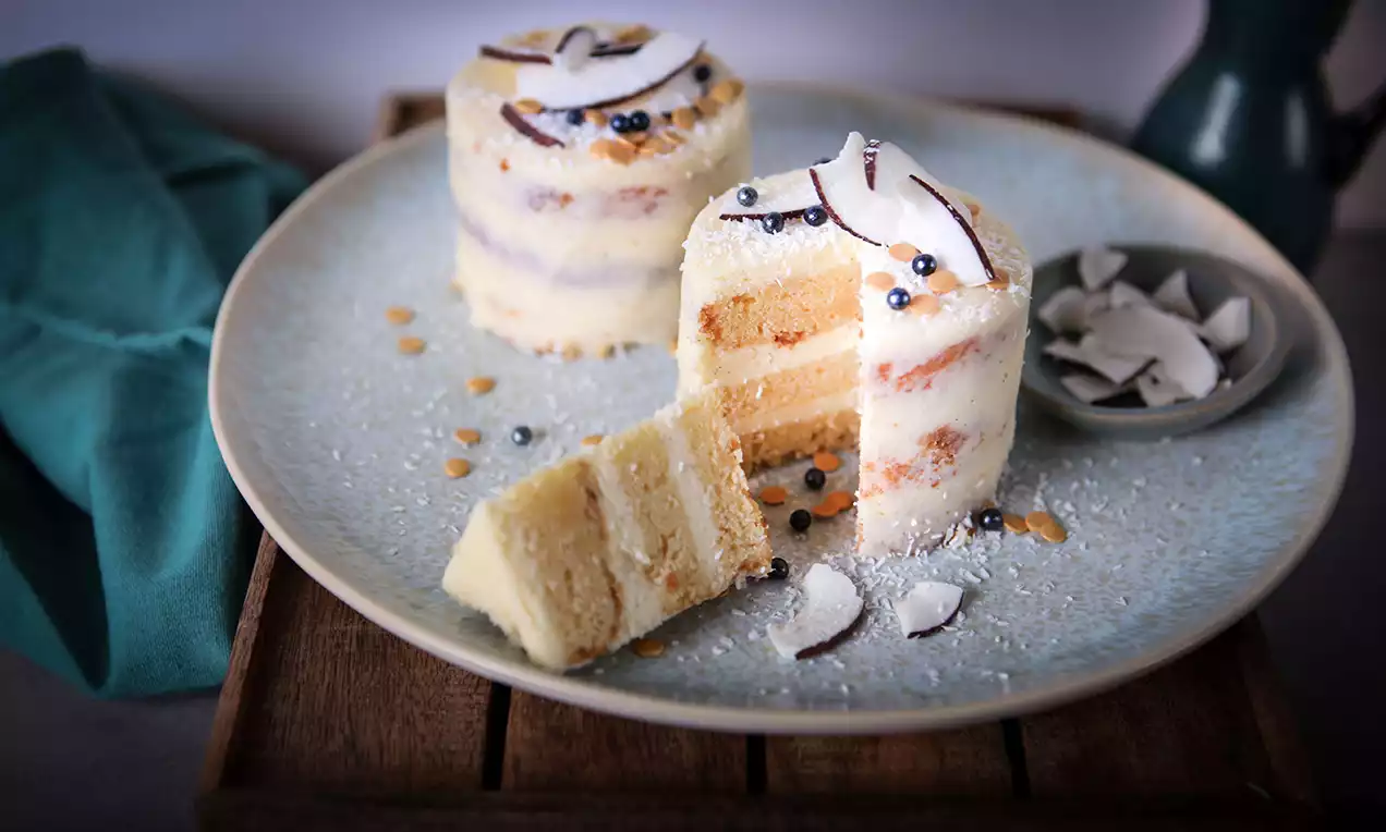 Zwei helle, schichtweise gefüllte Mini-Törtchen mit Kokosraspeln und silbernen Perlen auf einem großen Teller mit grünem Stoff drapiert