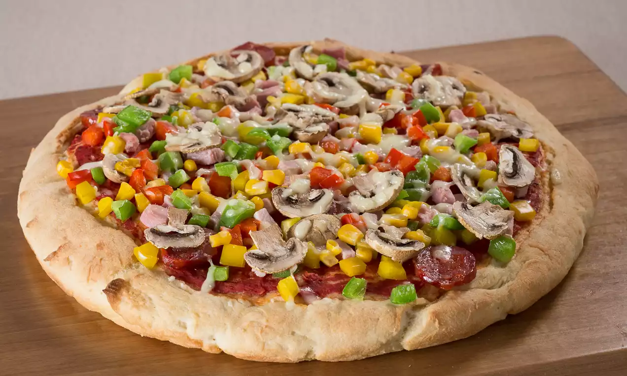 Ronde pizza met goudbruin gebakken korst, bedekt met kleurrijke stukjes paprika, maïs, champignons, hamblokjes en kaas op een houten plank
