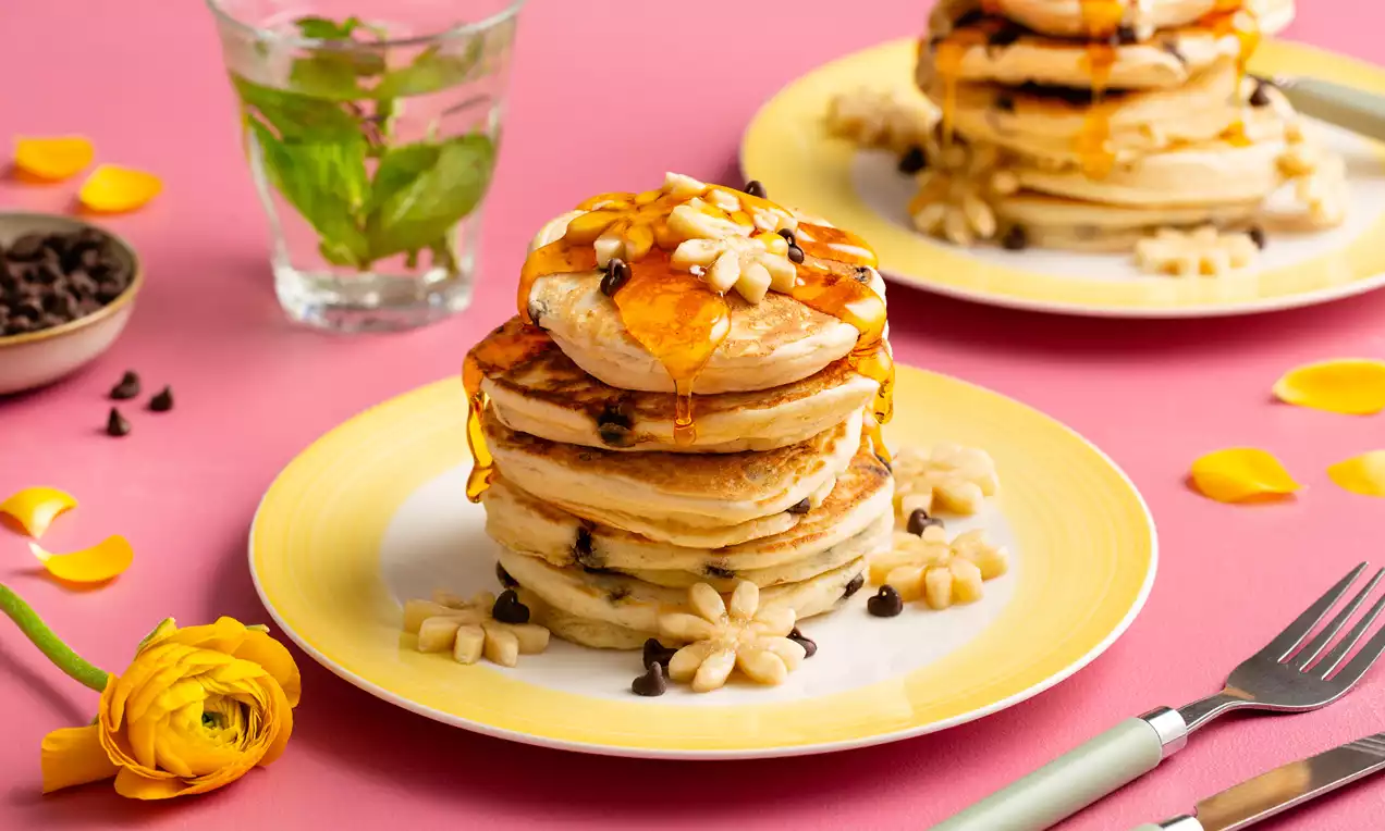 Stapels goudbruine American pancakes met chocolate chips en plakjes banaan, besprenkeld met honing op een lichtgeel bord