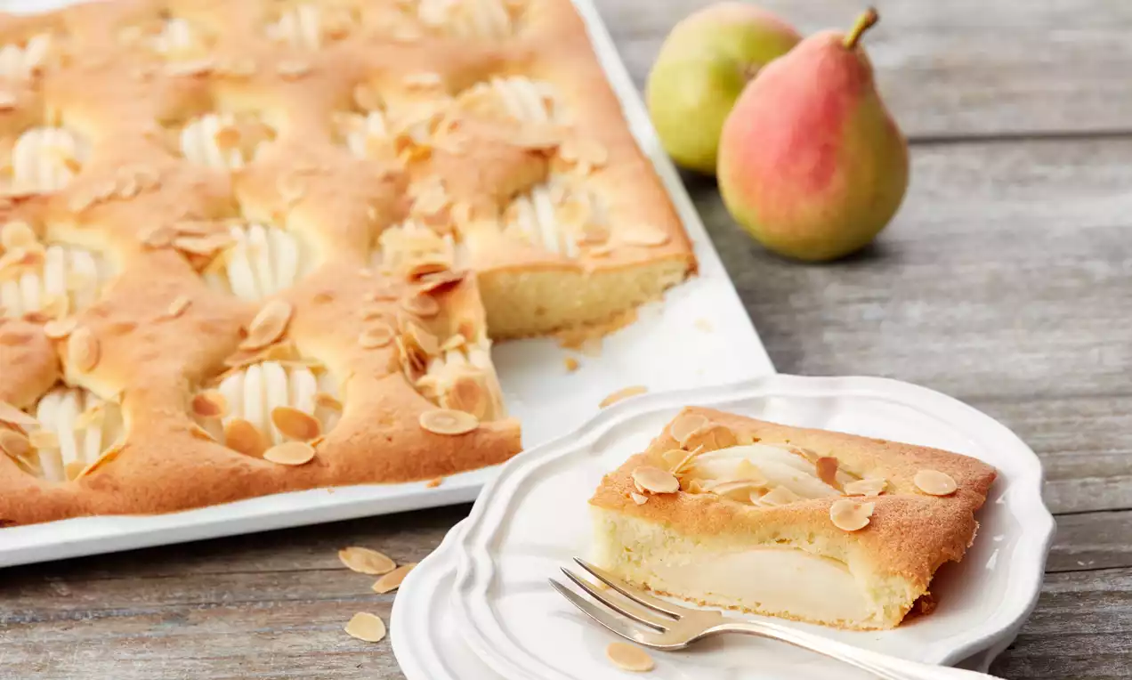 Birnenkuchen mit goldener Oberfläche, eingebetteten Birnenspalten und gerösteten Mandeln, ein Stück auf einem weißen Teller mit Gabel