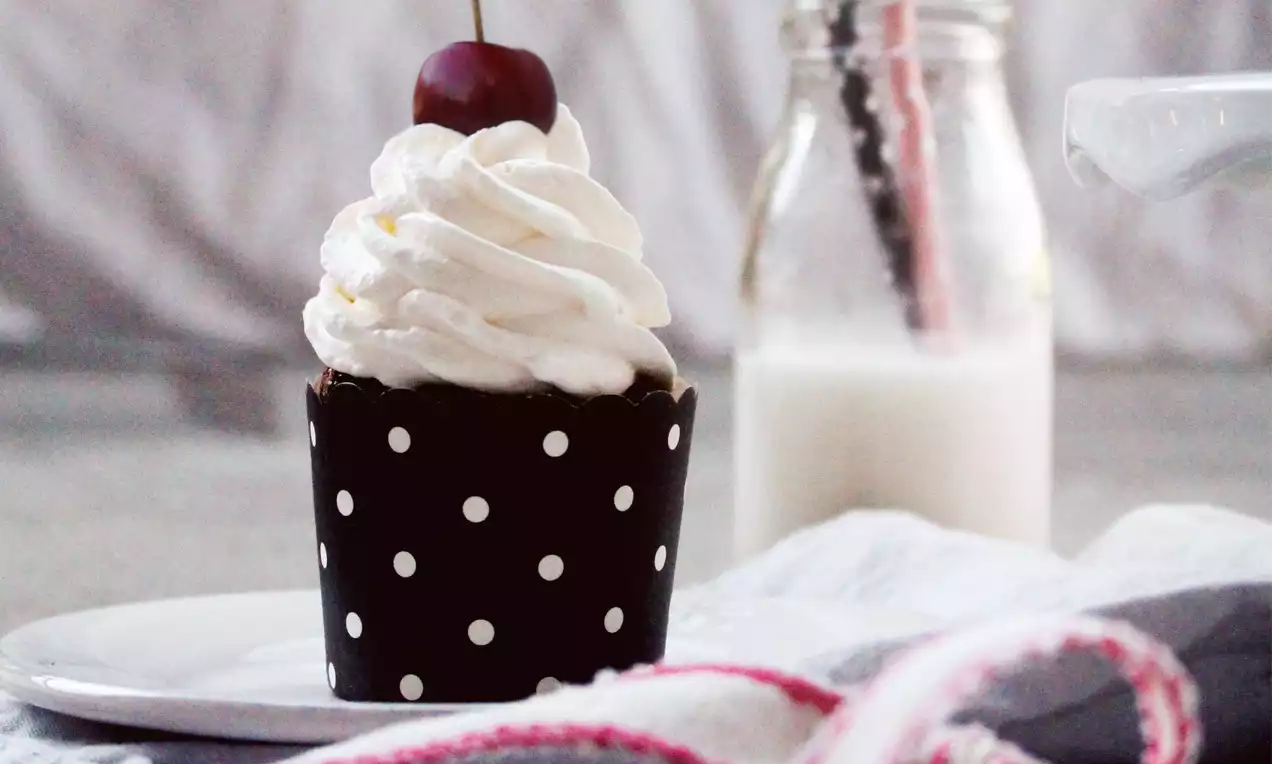 Kirsch-Cupcake im schwarz-weiß gepunkteten Förmchen mit spiralförmiger Sahnehaube und Kirsche, präsentiert auf weißem Teller mit Milchflasche