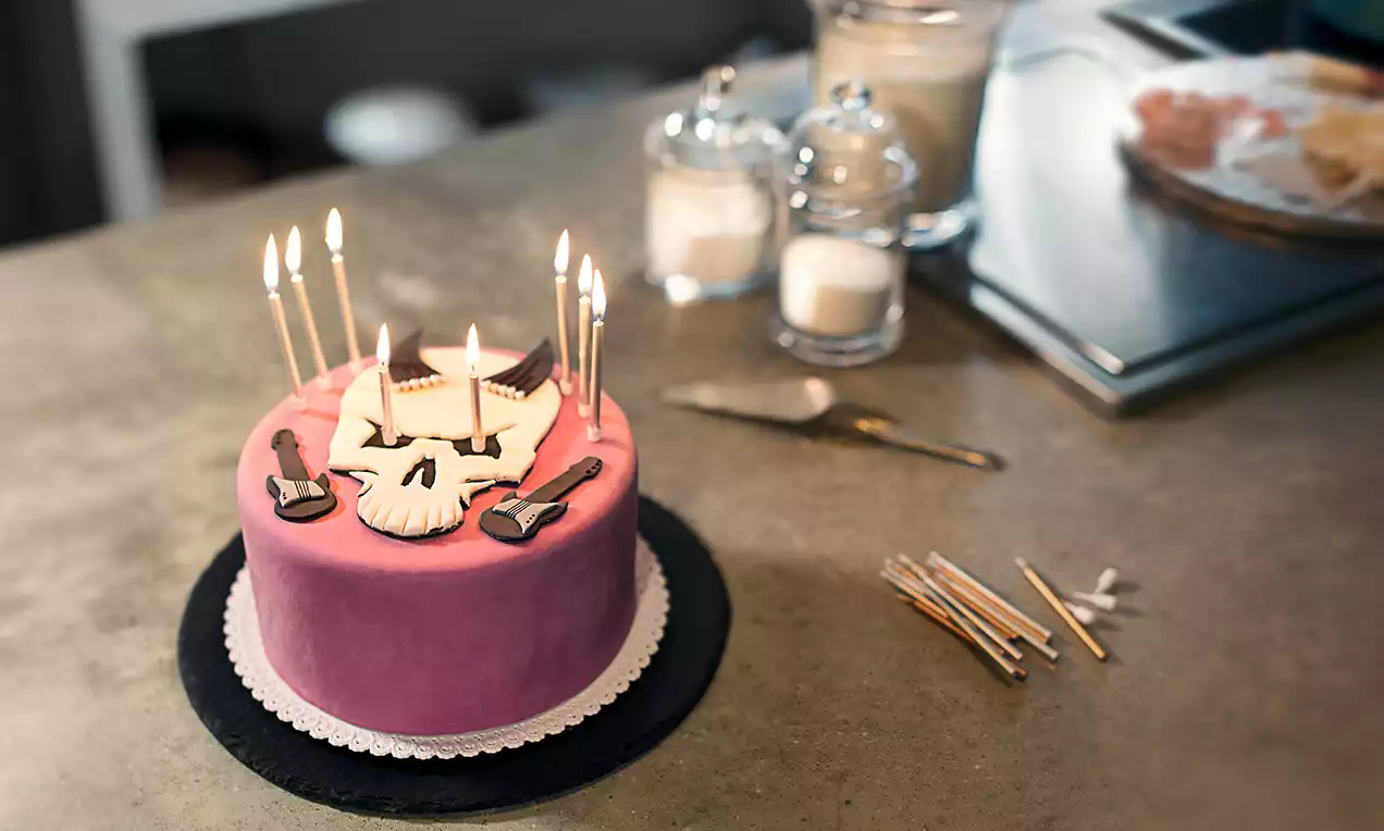 Runde Torte mit rosa Fondant, weißem Totenkopf und Zuckergitarren verziert, brennende Kerzen auf schwarzer Platte mit Spitzenrand