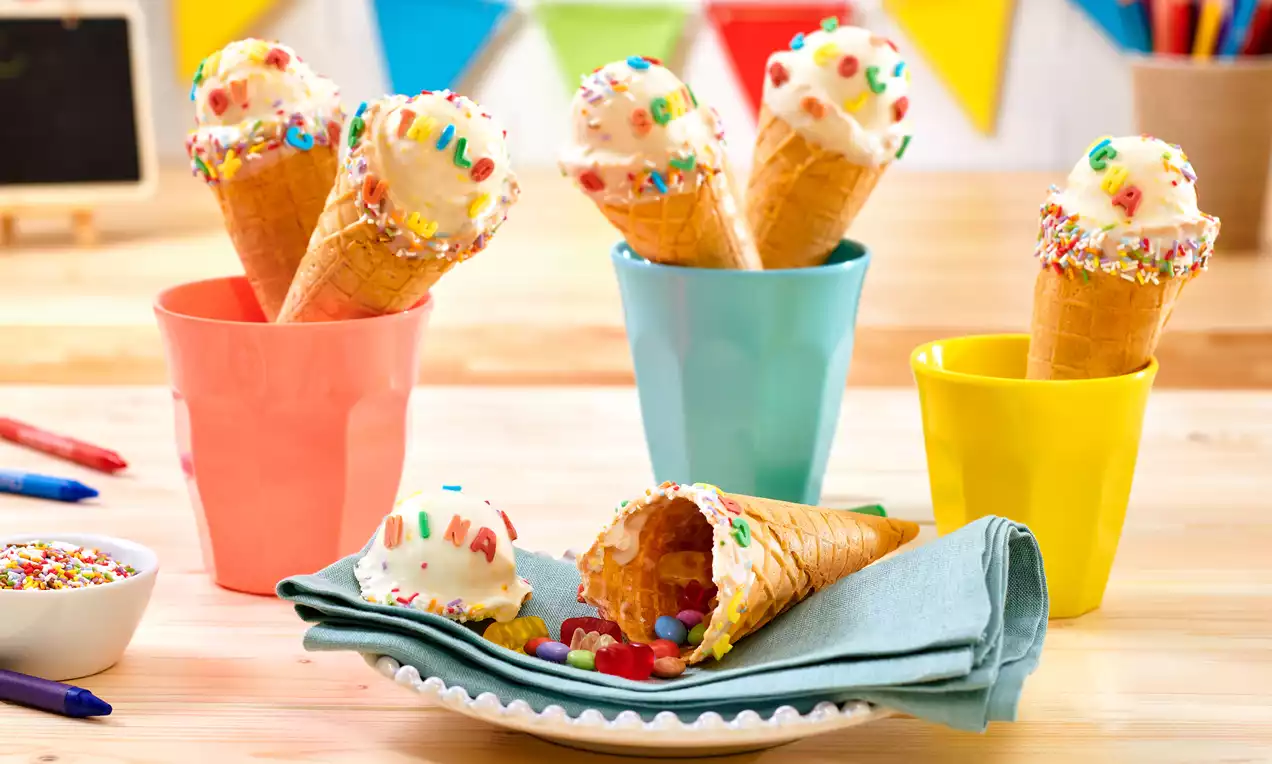 Eiswaffeln mit weißer Glasur und bunten Streuseln, gefüllt mit Süßigkeiten, arrangiert in pastellfarbigen Bechern und auf einem Teller mit Serviette