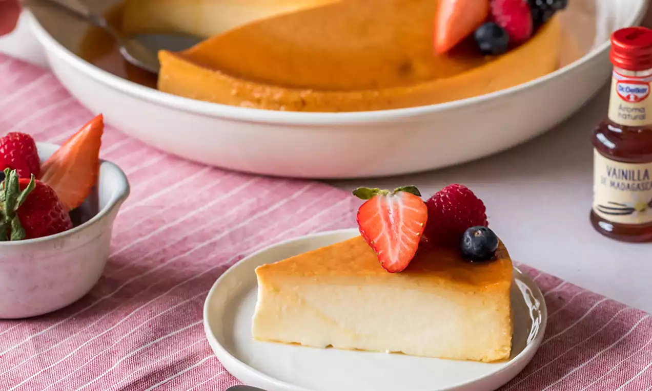 Porción triangular de flan de queso suave y dorado con textura lisa, decorada con fresas cortadas y arándanos sobre plato blanco