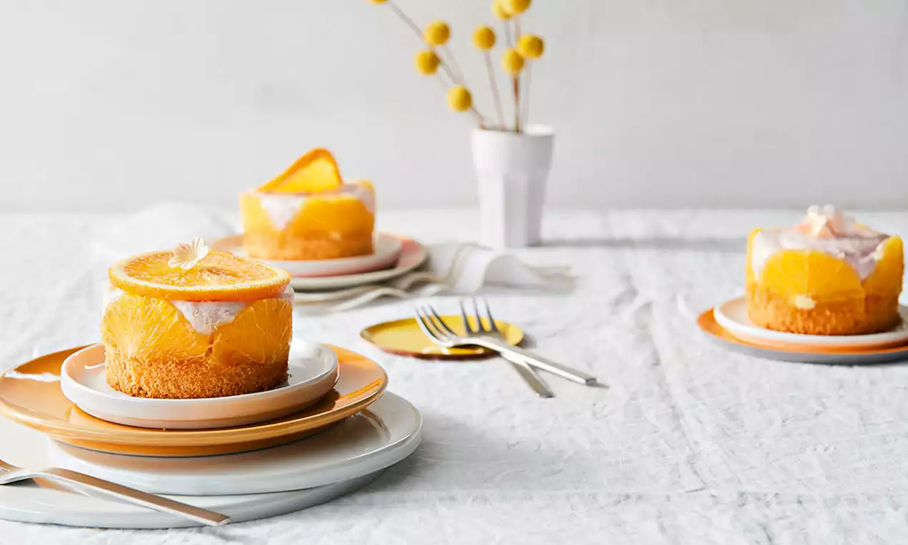 Tarte rotunde cu blat auriu, cremă fină de iaurt și felii translucide de portocale roșii, servite pe farfurii colorate pe o față de masă albă