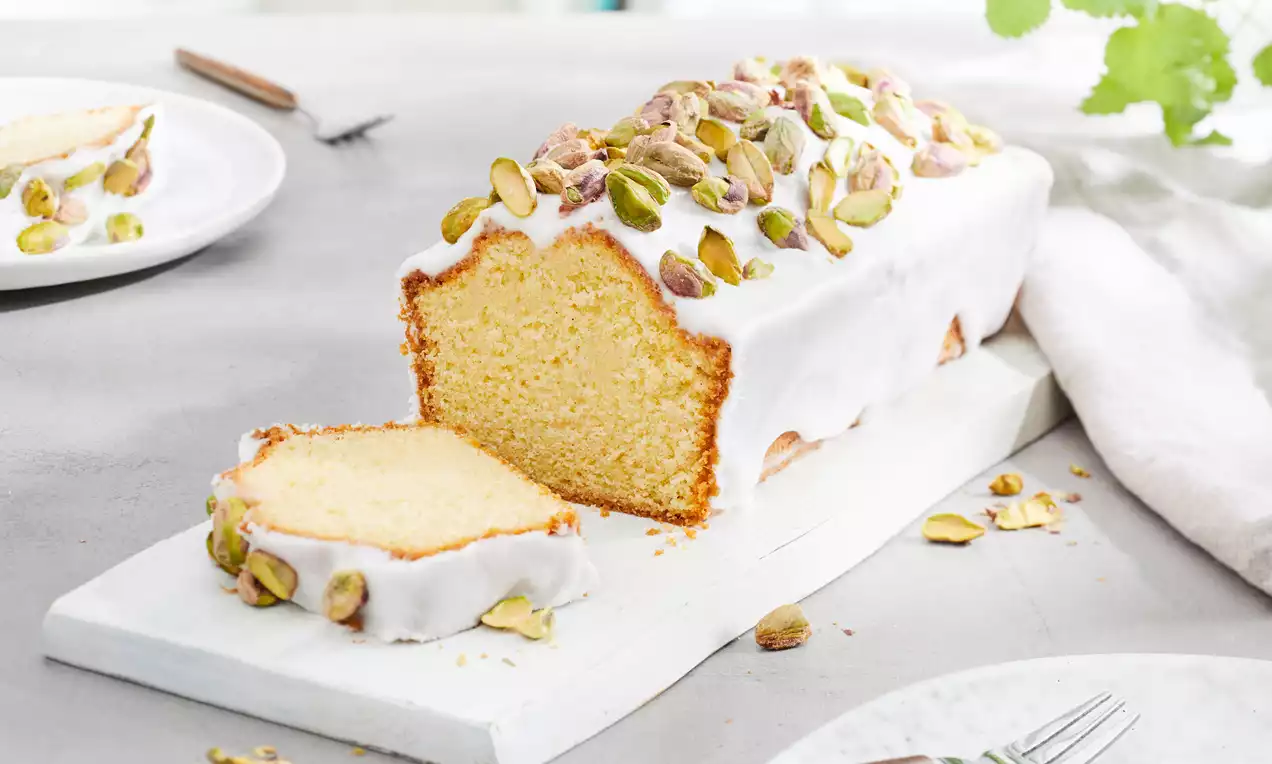 Langwerpige citroencake met witte glazuurlaag, bestrooid met groene pistachenoten, een plak afgesneden, liggend op een wit houten bord