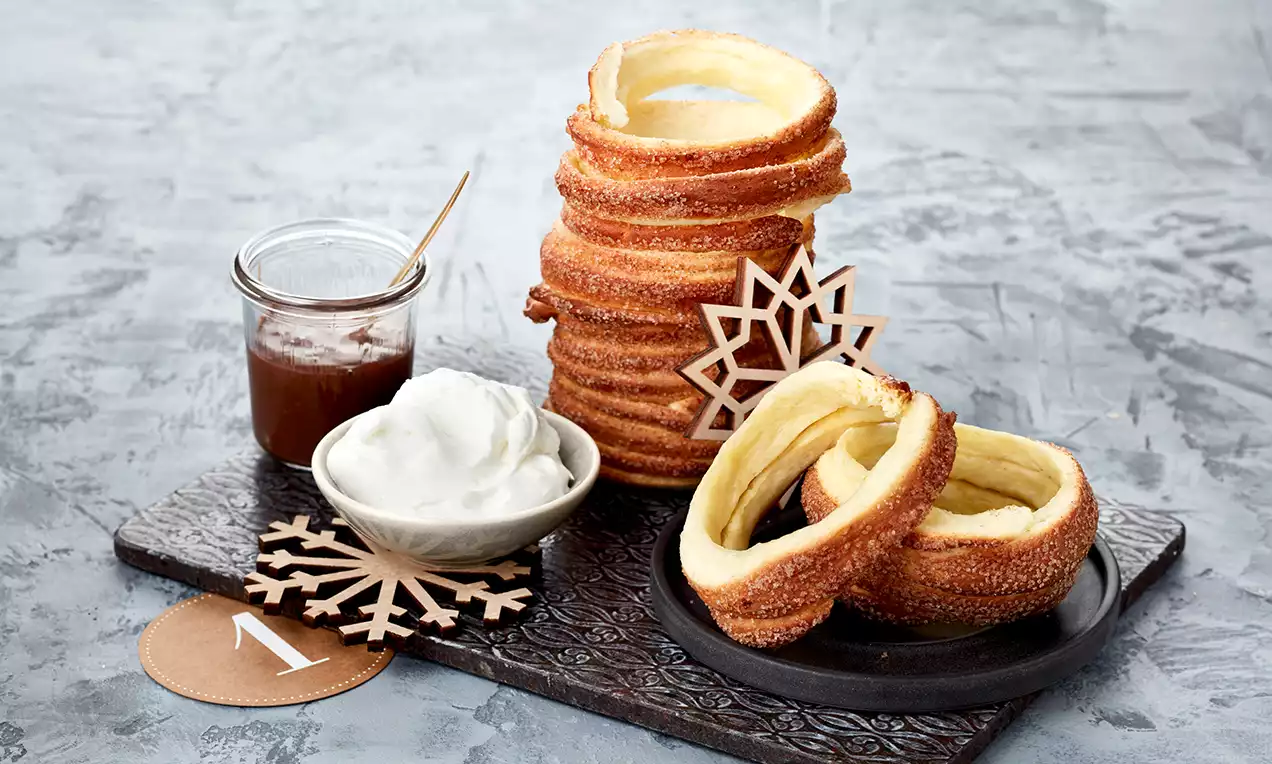 Goldbrauner Baumstriezel mit kristallinem Zuckermantel, turmartig gestapelt und auf einer Platte neben Sahne und Schokoladensauce arrangiert