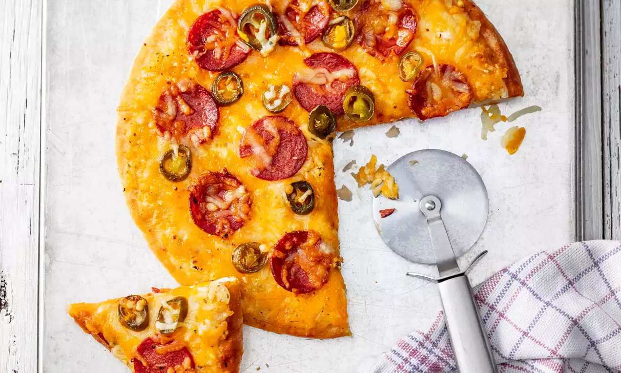 Goldbraune Pizza mit geschmolzenem Käse, roten Salamischeiben und grünen Jalapeños auf heller Oberfläche mit Pizzaschneider und Stofftuch