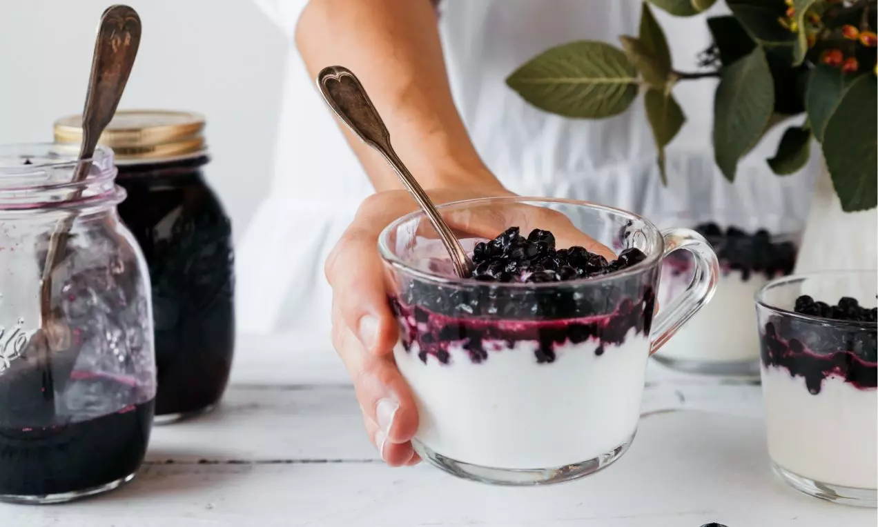 Číra sklenená miska plná bieleho jogurtu s vrstvou lesklých čučoriedok a tmavo fialového sirupu, podávaná s kovovou lyžičkou
