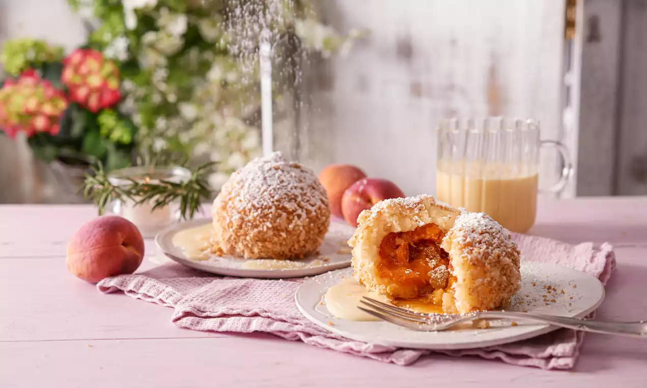 Goldbraune Aprikosenknödel mit knusprigen Bröseln und Staubzucker auf weißen Tellern, einer angeschnitten, serviert auf rosa Stofftuch