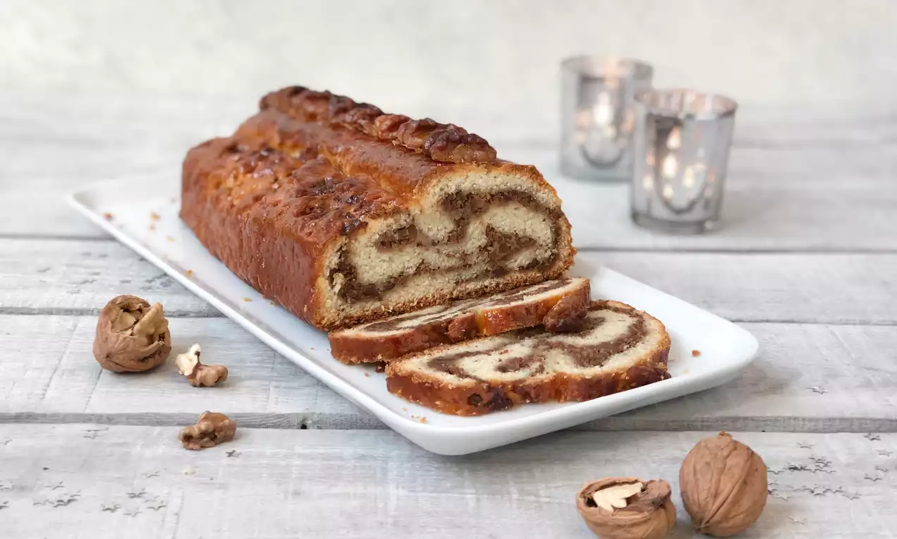 Walnuss-Wickelstollen mit goldbrauner Kruste, marmorierter Füllung und angeschnittenen Scheiben auf weißer Platte, umgeben von Walnüssen und Dekoration
