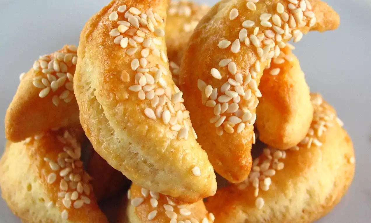 Altın rengi ve hafif kabarık yarım ay şekilli kurabiyeler, üzerleri bolca beyaz susamla kaplı, beyaz tabakta üst üste dizilmiş