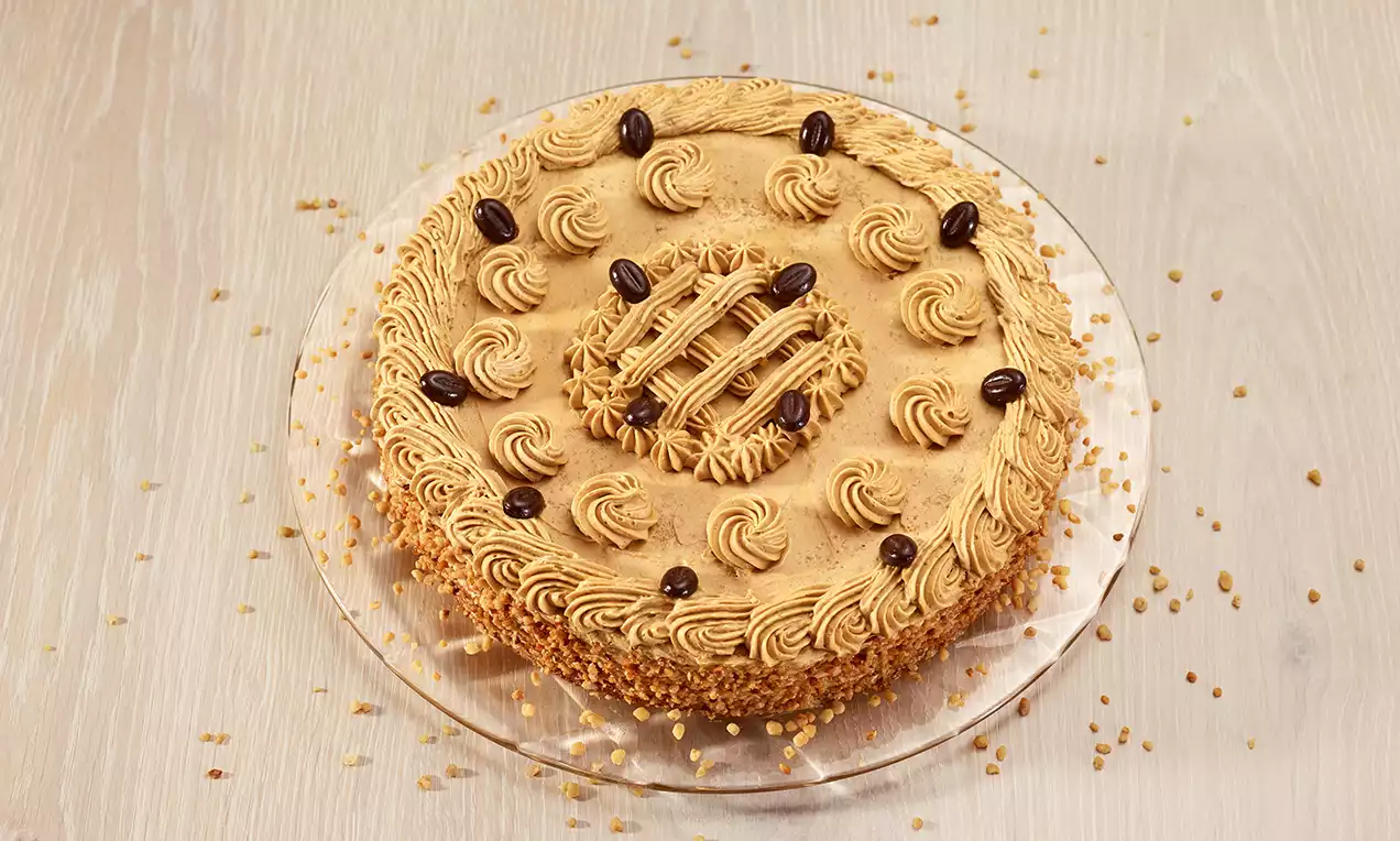 Torta al caffè rotonda con base marrone chiaro, riccioli di crema color nocciola, chicchi di caffè decorativi e bordo croccante di granella