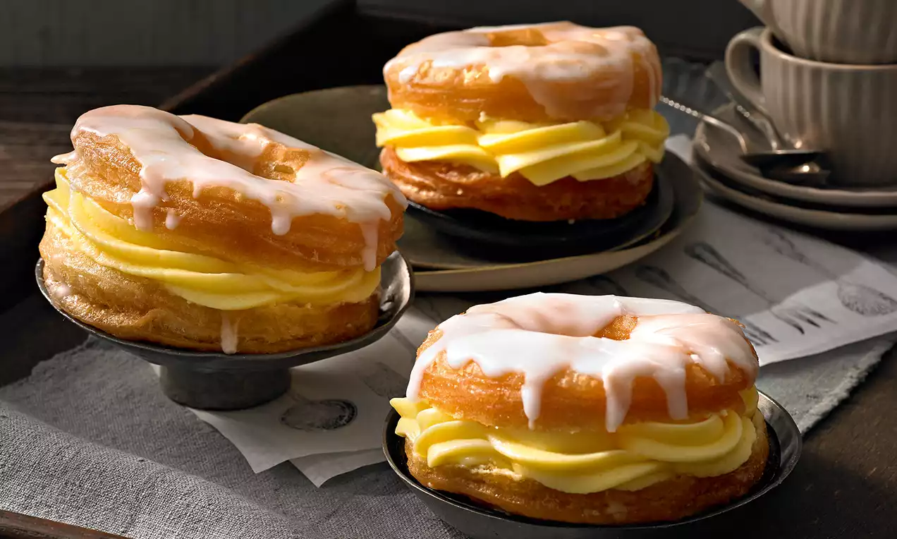 Zlatnosmeđi kroasani u obliku krofni s bijelom glazurom i slojem svijetložute kremaste puding kreme, posluženi na tamnim tanjirima