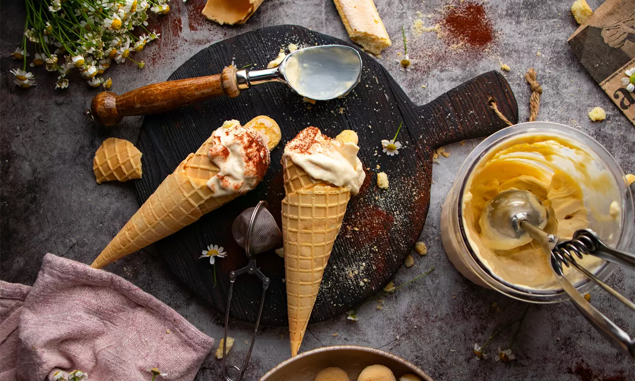 Два вафлени конуса, пълни с кремообразен сладолед тирамису, поръсен с какао, подредени върху тъмен дървен плот сред бишкоти и цветя