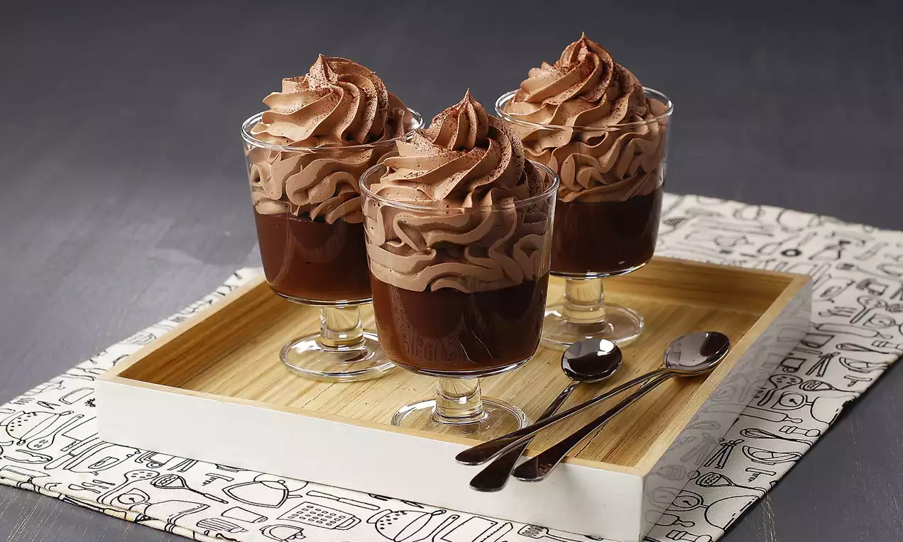 Ahşap tepsi üzerinde cam kupalarda koyu kahverengi puding ve üstünde sıkma kremalı, kıvrımlı çikolatalı mousse, yanında kaşıklarla düzenlenmiş
