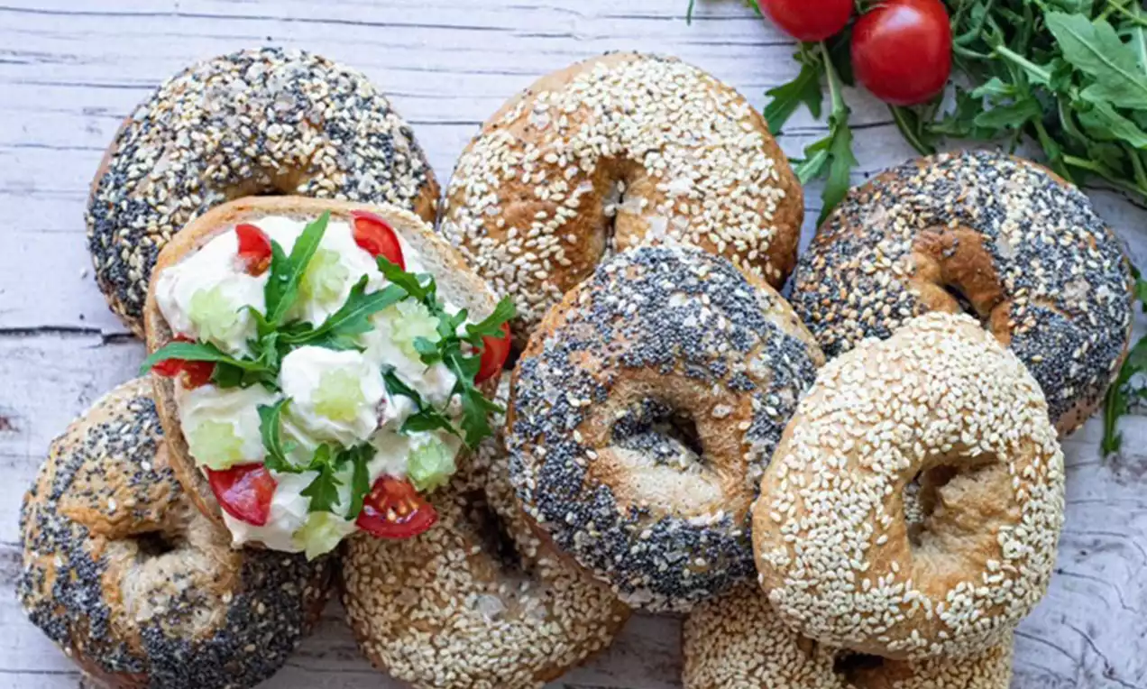 Runde Bagels in warmen Brauntönen, teils mit Mohn oder Sesam bedeckt, einer belegt mit Frischkäse, Tomaten, Gurken und Rucola, daneben frisches Gemüse
