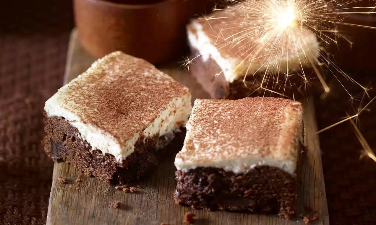 Drie vierkante brownies met een rijke bruine kleur, romige witte topping en cacaopoeder op een houten plank met een fonkelend sterretje