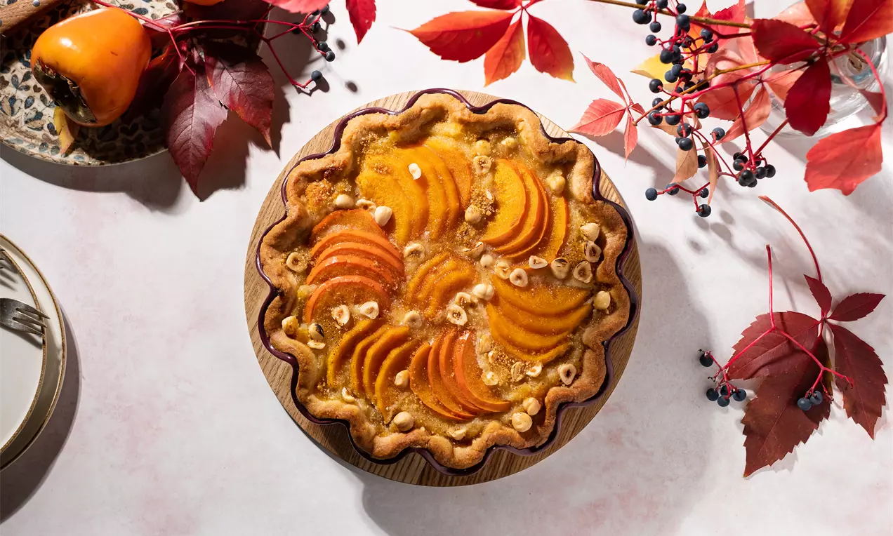 Тарт със златиста кора, подредени резени плодове с наситен оранжев цвят, хрупкави лешници и леко поръсени трохи, украсен с есенни листа